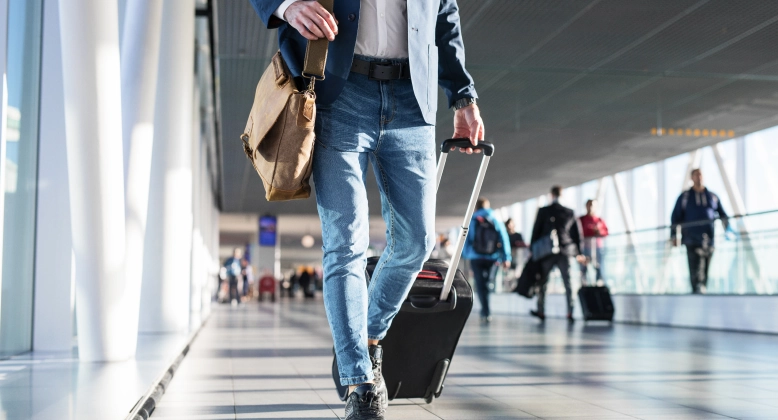 Below-the-waist photo of a man walking through an airport with a suitcase and satchel.